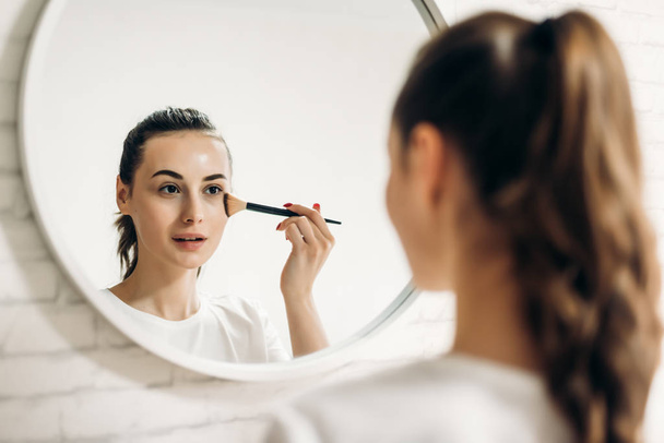 Beautiful young woman applying makeup in bathroom. - Фото, изображение