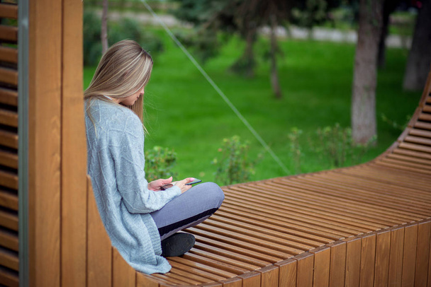 Chica rubia sentada con las piernas en un banco y mirando un teléfono inteligente, vista desde la parte posterior
 - Foto, Imagen