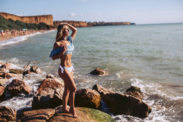 Una bella ragazza snella si trova su una spiaggia rocciosa guardando in lontananza. Caldo giorno di riposo estivo
. - Foto, immagini