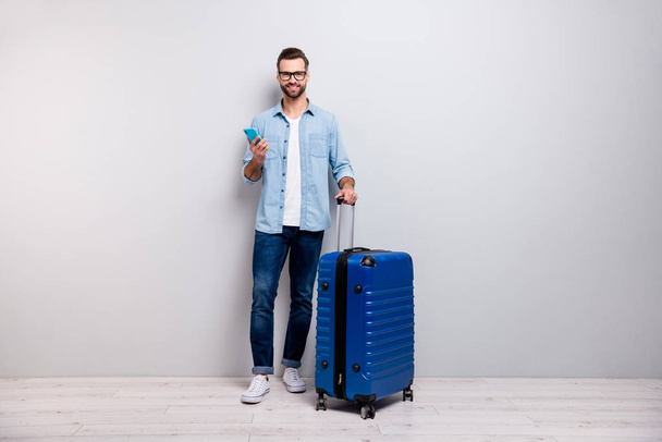 Full length photo of handsome hipster guy holding telephone big tourist bag have student visa europe traveler wear specs denim outfit isolated grey color background - 写真・画像