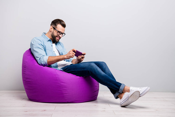 Full length profile photo of positive guy sitting comfy violet armchair holding telephone playing new game wear specs casual denim outfit isolated grey color background - Fotografie, Obrázek