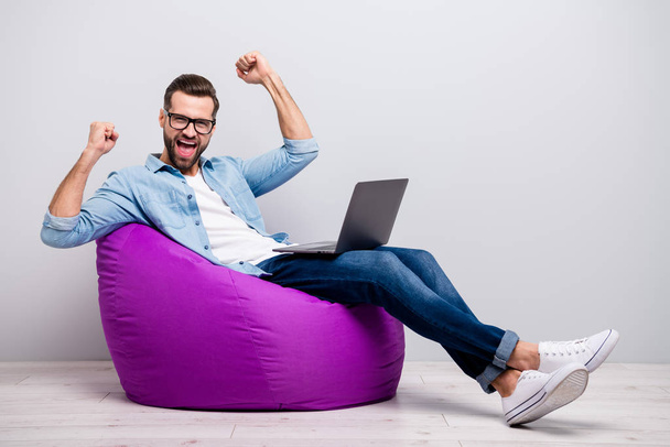 Full size photo of crazy guy hold notebook sitting comfortable soft violet armchair celebrating startup success freelancer wear specs casual denim outfit isolated grey color background - Foto, afbeelding