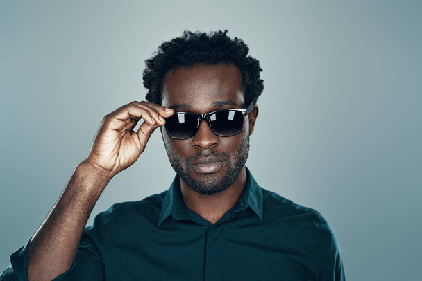 Handsome young African man looking at camera and adjusting eyewear while standing against grey background - Zdjęcie, obraz