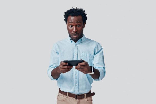 Komea nuori afrikkalainen mies pelaa mobiilipelejä samalla kun seisoo harmaata taustaa vasten
 - Valokuva, kuva