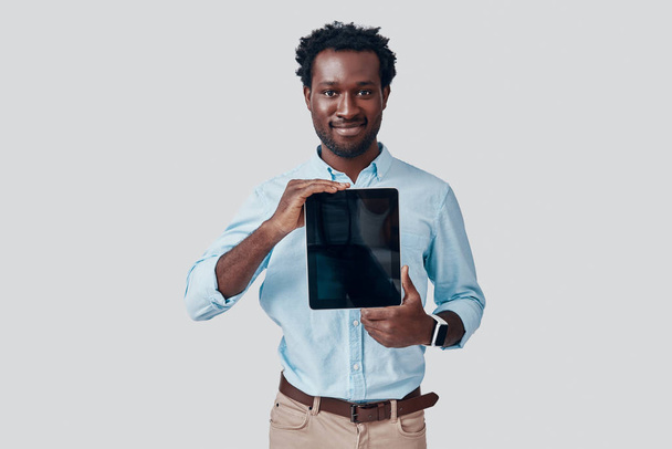 Przystojny młody Afrykanin wskazujący miejsce na cyfrowym tablecie i uśmiechnięty stojąc na szarym tle - Zdjęcie, obraz