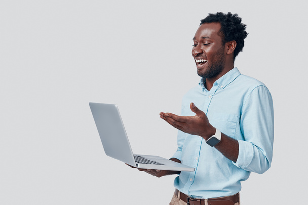 Hombre africano joven guapo usando el ordenador portátil y sonriendo mientras está de pie sobre fondo gris
 - Foto, Imagen
