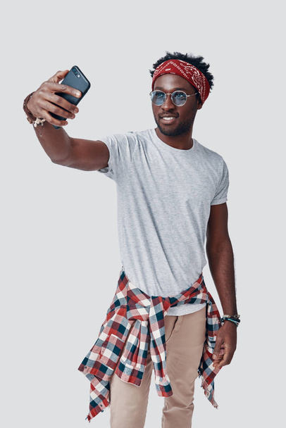 Όμορφος νεαρός Αφρικανός βγάζει σέλφι και χαμογελάει ενώ στέκεται στο γκρίζο φόντο - Φωτογραφία, εικόνα