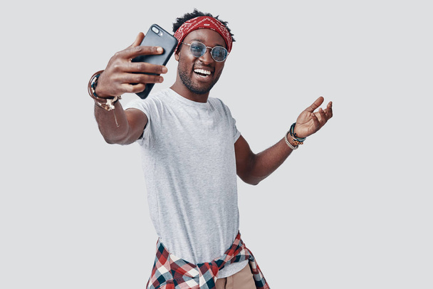 Όμορφος νεαρός Αφρικανός βγάζει σέλφι και χαμογελάει ενώ στέκεται στο γκρίζο φόντο - Φωτογραφία, εικόνα