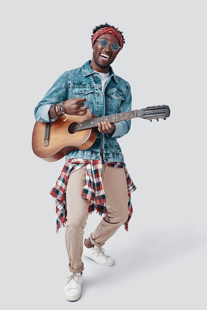 Longitud completa de guapo joven africano tocando la guitarra y sonriendo mientras está de pie sobre fondo gris
 - Foto, imagen