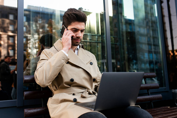 Young casual businessman in trench coat confidently working on laptop and talking on cellphone on city street - Fotografie, Obrázek