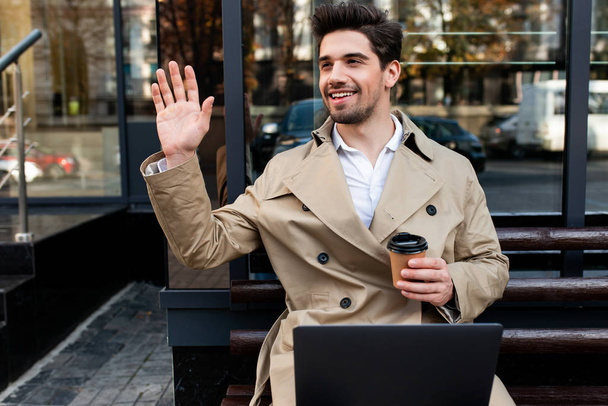 Young attractive smiling man in trench coat with laptop and coffee joyfully waving hand hello on city street - Photo, Image