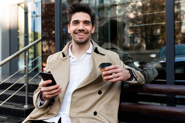 Jovem homem casual positivo em casaco de trincheira com café para ir e celular olhando alegremente na câmera na rua da cidade
 - Foto, Imagem