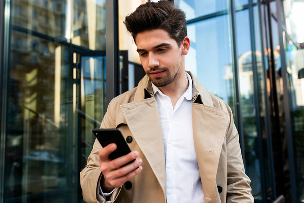Молодой стильный мужчина в пальто с мобильником на городской улице
 - Фото, изображение