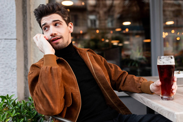 Young attractive casual businessman talking on cellphone resting with beer in cafe on street - Photo, image