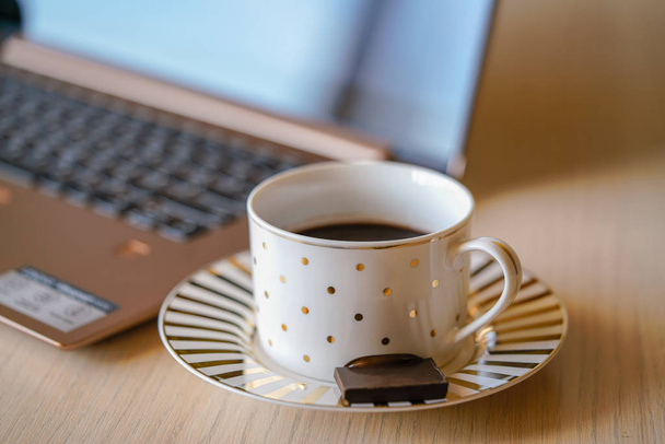 Чашка кофе на деревянном столе с ноутбуком на заднем плане. Кусок черного шоколада на блюдце, перерыв в офисе для освежения
 - Фото, изображение