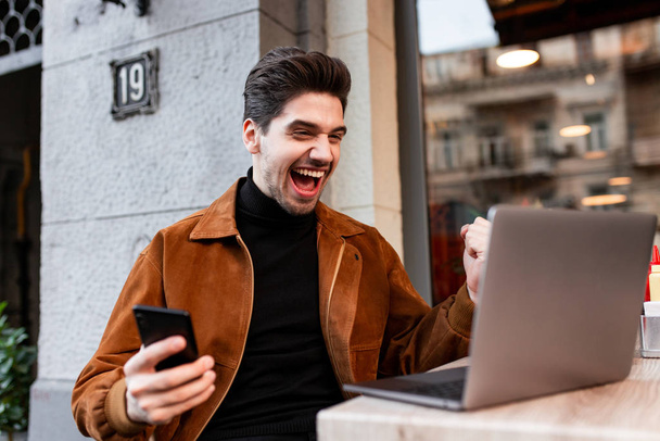 Giovane allegro uomo casual che lavora sul computer portatile con il cellulare e gioire in caffè sulla strada
 - Foto, immagini