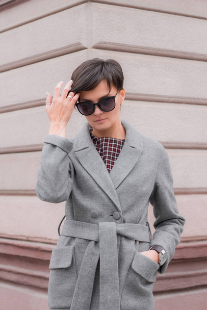 Autumn fashion outdoor. The brunette girl with short hair in fashionable stylish grey coat and sunglasses, posing on the background of building. Street fashion.Autumnal lifestyle - Fotografie, Obrázek