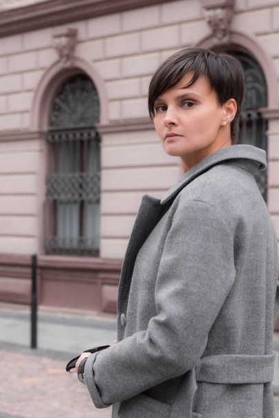 Autumn fashion outdoor. The brunette girl with short hair in fashionable stylish grey coat, posing on the background of building. Street fashion.Autumnal lifestyle - Fotó, kép