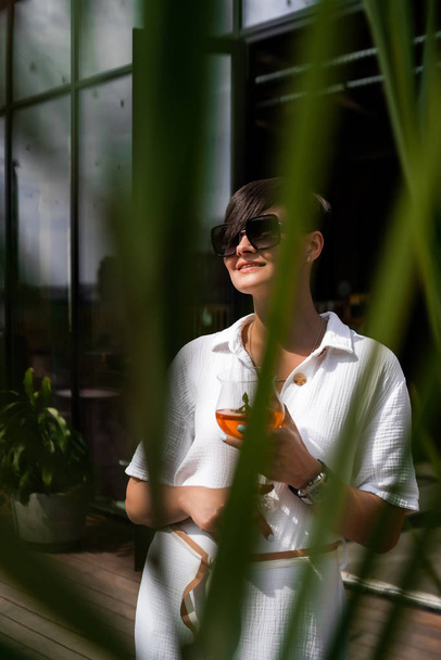 Beautiful woman with short brunette hair in white clothes and sunglasses. Fashion street photography. Fashion model posing with aperol cocktail on wood background. - Φωτογραφία, εικόνα