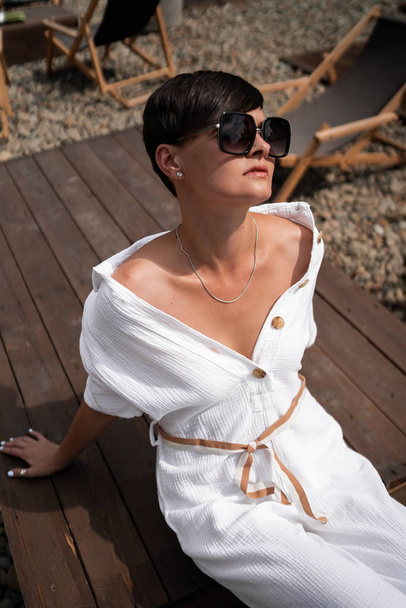 Beautiful woman with short brunette hair in white clothes and sunglasses. Fashion street photography. Fashion model is sitting and relax on brown wood background. sunglasses shop - Fotografie, Obrázek