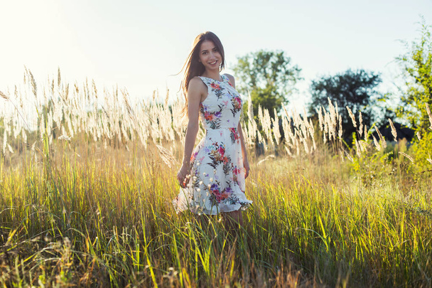 Menina atraente no vestido midi floral branco no jardim de verão com pose de grama longa na luz do pôr do sol
 - Foto, Imagem