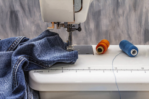 Sewing machine foot on denim. Scissors, spools of thread. Stands on a table, wooden backdrop - Фото, изображение