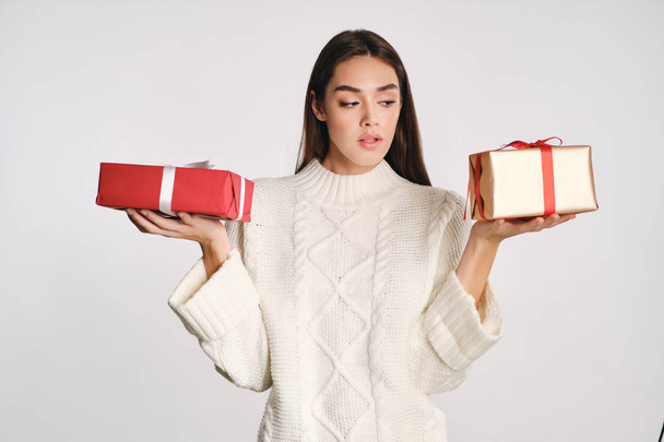 Beautiful girl in cozy sweater thoughtfully holding gift boxes in hands over white background - Photo, Image