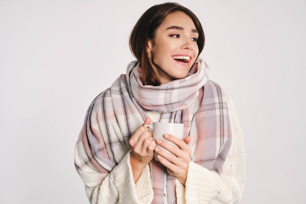 Attractive cheerful girl with cozy scarf holding cup with warm drink happily looking away over white background - Foto, afbeelding