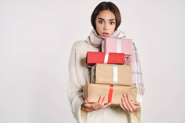 Attractive shocked girl in sweater with scarf holding gift boxes amazedly looking in camera over white background - Photo, Image