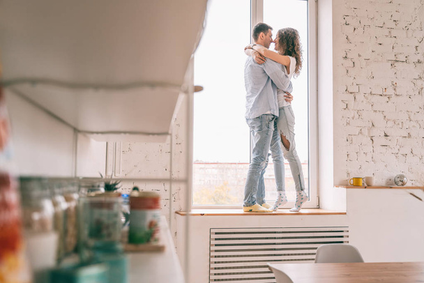 Young happy couple hugging on the windowsill in their apartment, - 写真・画像