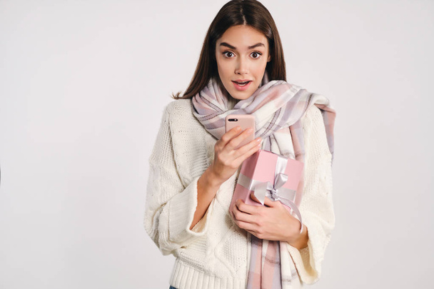 attraktives Mädchen in kuscheligem Pullover mit Schal, der Geschenkbox und Handy hält, blickt überraschend in die Kamera über weißem Hintergrund - Foto, Bild
