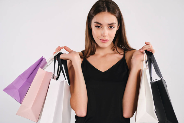 Gyönyörű elegáns lány fekete ruhában gazdaság bevásárló táskák magabiztosan keres a kamera felett fehér háttér - Fotó, kép