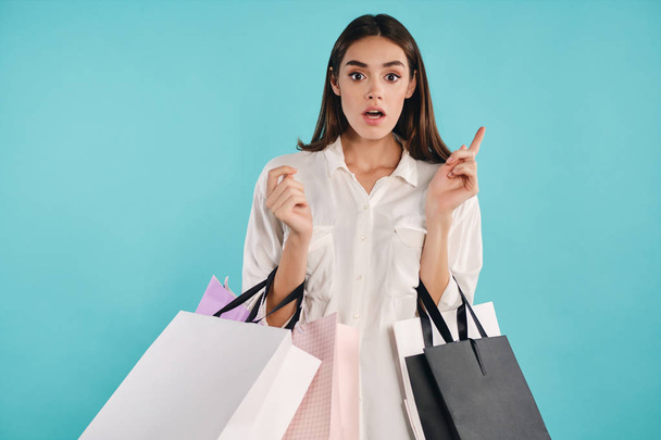 Attractive casual girl with shopping bags amazedly looking in camera having idea over colorful background - Foto, Imagen