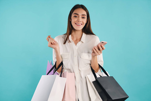 Pretty casual girl with shopping bags and cellphone happily looking in camera over colorful background - Foto, Imagen