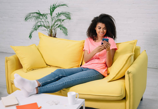 Νεαρή ελκυστική Αφροαμερικανή γυναίκα που κατέχει smartphone, επικοινωνία, παραγγελία τροφίμων στην ιστοσελίδα, κάθεται σε άνετο κίτρινο καναπέ. Ευτυχισμένο κορίτσι που χρησιμοποιεί το κινητό app για online αγορές, μείνετε στο σπίτι - Φωτογραφία, εικόνα