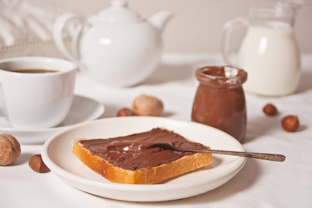 Bread toast with chocolate cream butter, jar of chocolate cream, cuop of tea, jar of milk, teapot on the white background. - Foto, Imagem