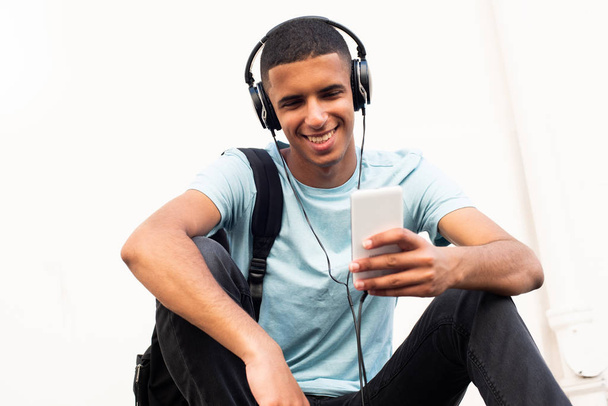 Retrato de um jovem norte-africano moderno sentado na parede branca ouvindo música com telefone celular e fones de ouvido
 - Foto, Imagem