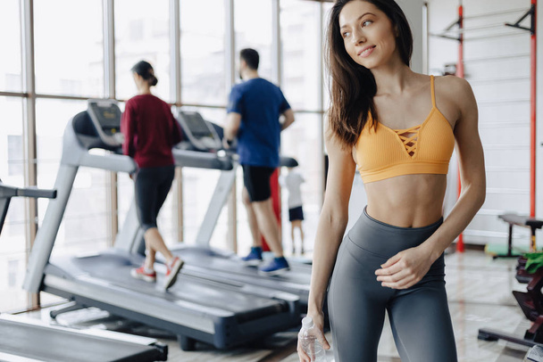 young attractive girl in the gym drinking water on the background of treadmills - Photo, Image