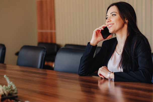 Молодая привлекательная эмоциональная девушка в деловой одежде сидит за столом с телефоном в офисе или аудитории
 - Фото, изображение