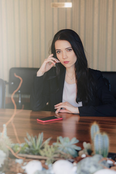 Young attractive emotional girl in business style clothes sitting at desk with phone in office or audience - Фото, зображення