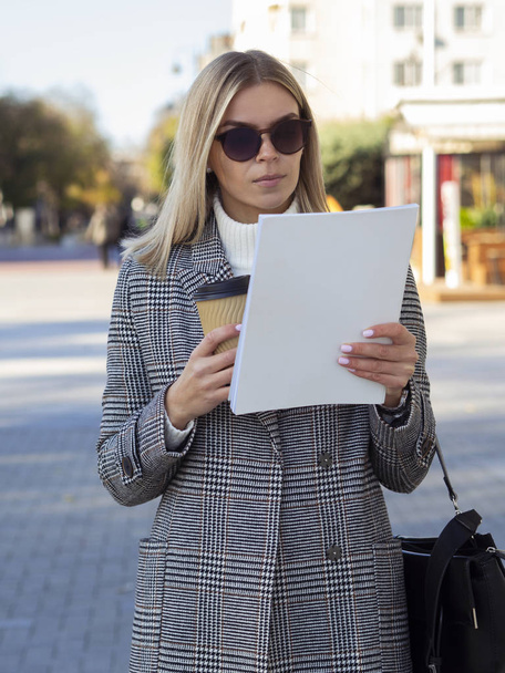 White European girl in sunglasses and a coat on a cold spring day holds a cup of coffee and reading a white magazine with blank cover with copy space for your mockup - Photo, Image