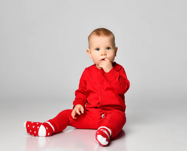 Little toddler boy in red infant bodysuit is sitting looking puzzled with free text space on gray full body - Foto, Imagem