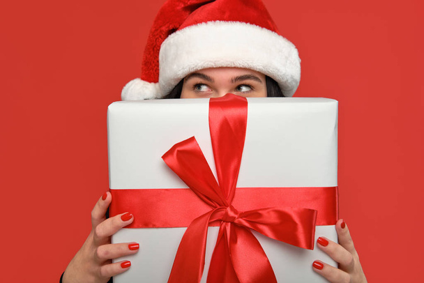 Woman in a black dress and Santa hat hiding behind Christmas giftbox of white color with red ribbon. - Φωτογραφία, εικόνα