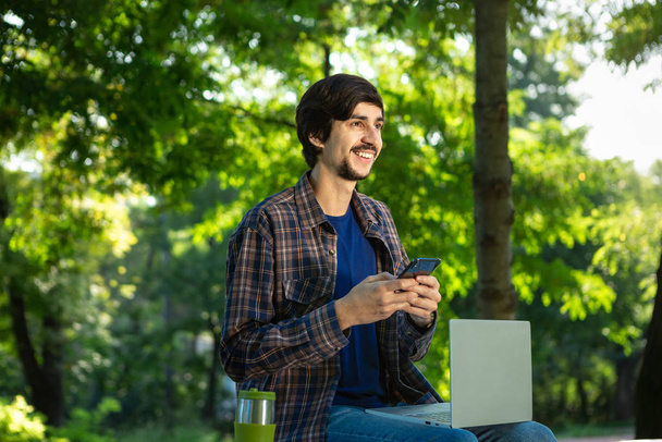 Młody wolny strzelec z brodą i wąsami siedzi z laptopem w parku i trzyma smartfona. - Zdjęcie, obraz