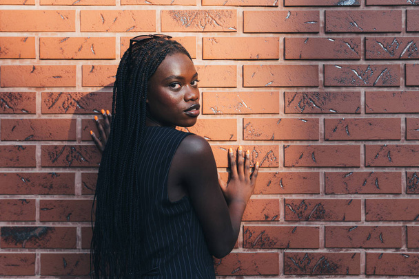 Attractive afroamarican girl in black striped vest stands back to the camera and looks at the camera over her shoulder holds her hands on the wall, red brick wall on the background - Photo, Image