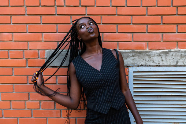 Νεαρή Αφρο-Αμερικανίδα με μαύρα ριγέ ρούχα κρατά τα μαλλιά της στο χέρι και κοιτάζει την πάνω αριστερή γωνία. - Φωτογραφία, εικόνα