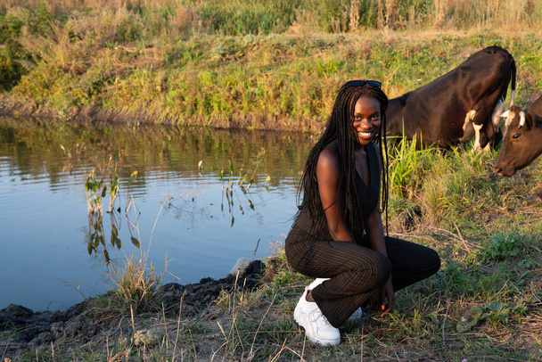 Счастливая улыбающаяся африканская девушка в черной одежде присела и, глядя в камеру, корова пьет воду из озера на заднем плане.
 - Фото, изображение