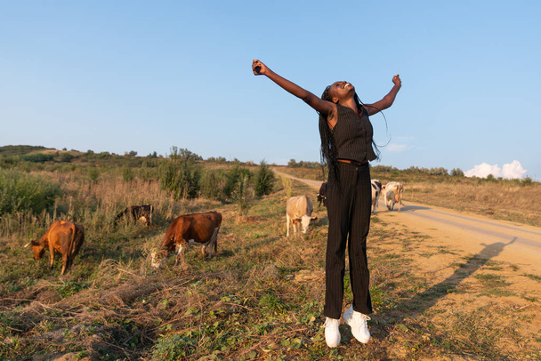 Fiatal boldog mosolygós afrikai lány fekete ruhában ugrik felemelt kézzel a mezőn, tehenek legeltek a háttérben - Fotó, kép