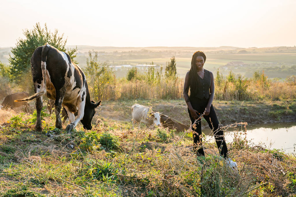 Jeune fille africaine en vêtements noirs se tient parmi les champs, les vaches boivent de l'eau du lac sur le fond
 - Photo, image