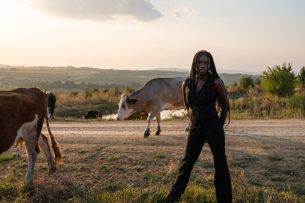Fiatal afrikai lány fekete ruhában áll a mezőn, a kezét a háta mögött tartja, tehenek legelnek a háttérben. - Fotó, kép